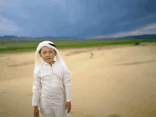 a young boy wearing a white shirt and a white head scarf stands in a desert