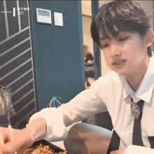 a young man in a white shirt and tie is sitting at a table eating food with hybe written on the wall behind him