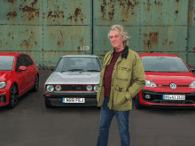 a man in a green jacket stands in front of a row of cars one of which has a license plate that says ag51fej