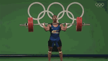 a man is squatting down while holding a barbell over his head with the olympic rings in the background .