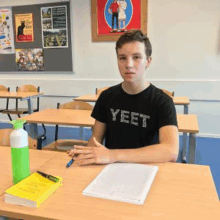 a young boy is sitting at a desk in a classroom wearing a black shirt that says yeet .