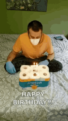 a man wearing a mask and gloves is sitting on a bed holding a birthday cake made out of toilet paper rolls .