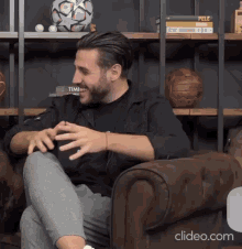 a man in a black shirt is sitting in a chair in front of a bookshelf with soccer balls on it