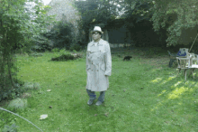 a man in a trench coat and hat stands in a grassy area