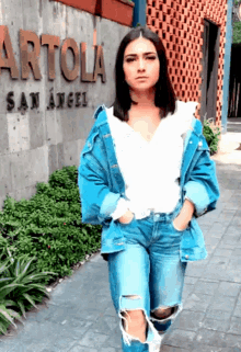 a woman wearing ripped jeans and a denim jacket stands in front of a sign that says artola san angel