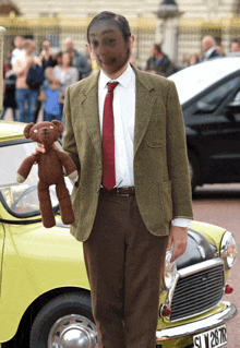 a man dressed as mr bean is holding a teddy bear in front of a yellow car with license plate slw 2876