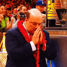 a man in a suit and tie with a red lei around his neck is praying in front of a crowd .