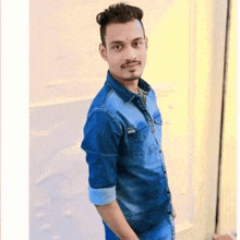 a young man wearing a blue denim shirt and blue jeans is standing in front of a white wall .