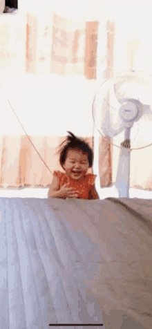 a little girl is sitting on a bed with a fan in the background .