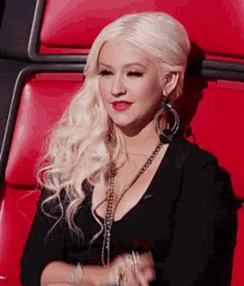 a blonde woman is sitting in a red chair wearing a black top and earrings .