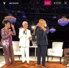 three women singing in front of microphones on a stage with a live button in the corner