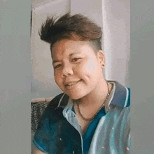 a young man with short hair is smiling for the camera while wearing a blue shirt and a necklace .