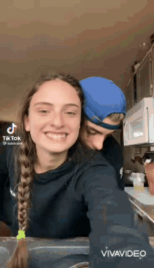 a man and a woman are posing for a picture in a kitchen and the woman is wearing a braided ponytail .