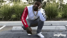 a man is squatting down in a parking lot while wearing a white shirt with arabic writing on the sleeves .