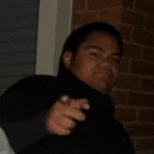 a man in a black jacket is pointing at the camera while standing in front of a brick wall .