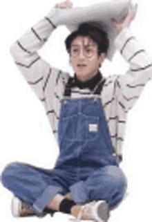 a young man in overalls is sitting on the floor holding a pillow on his head .