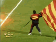 a man in an adidas jacket is holding a flag on a soccer field with smoke coming out of it .