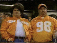 two men wearing orange jackets with the number 98 on them are standing next to each other .