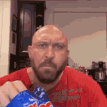 a bald man with a beard is holding a bag of chips and looking at the camera .