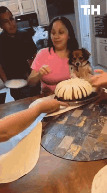 a woman in a pink shirt is holding a dog in her lap while a person holds a cake with the letters th above it