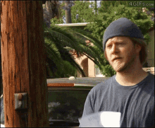 a man wearing a beanie and a t-shirt is standing in front of a palm tree and a car ..