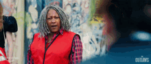 a woman wearing a red vest and plaid shirt is standing in front of a wall with graffiti on it .