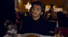 a man in a black suit sits at a table with a plate of food and a glass of water