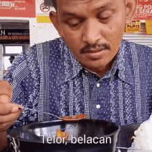 a man in a blue and white plaid shirt is eating from a black bowl with the words telor belacan written on the bottom