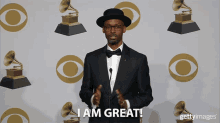 a man in a suit and bow tie stands in front of a wall of grammy awards and says i am great