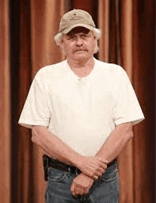 an older man wearing a hat and a white shirt is standing in front of a curtain .