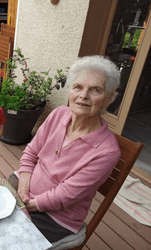 an elderly woman in a pink sweater sits at a table outside