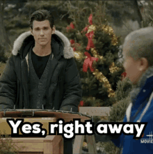 a man in a green jacket stands in front of a christmas tree while a woman says " yes right away "