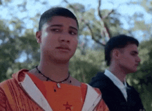 two young men are standing next to each other in a park . one of the men is wearing a necklace .