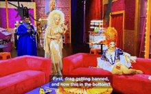 a drag queen is sitting on a red couch and talking to another drag queen