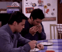 two men are sitting at a table eating food in front of a refrigerator that has a picture of a group of people on it