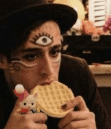 a man with an eye painted on his face is eating a waffle with a stuffed animal in his hand .