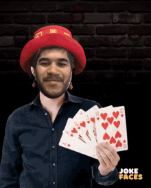 a man wearing a red hat is holding four playing cards in front of a brick wall with joke faces written on it