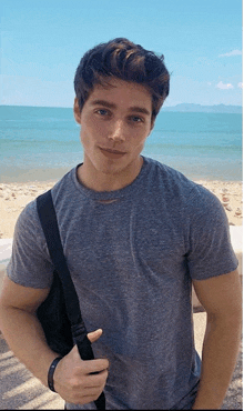 a young man wearing a grey t-shirt and a black bag stands in front of the ocean