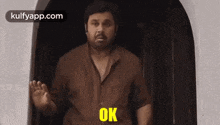 a man in a brown shirt is standing in front of a door and saying `` ok '' .