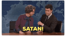 a man and a woman are sitting at a table and the woman is saying satan