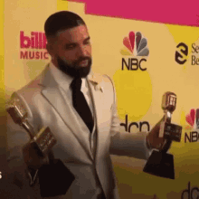 a man in a suit and tie is holding two trophies in front of a wall that says bill music and nbc