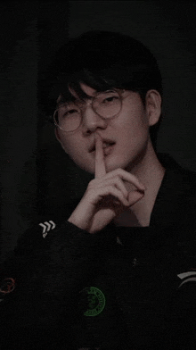 a young man wearing glasses and a black jacket is holding his finger to his lips .