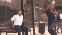 a man in a white shirt is sitting on a bench while a man in a blue shirt jumps in front of him