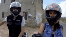 a man and a woman wearing motorcycle helmets are standing in front of a building that has graffiti on it