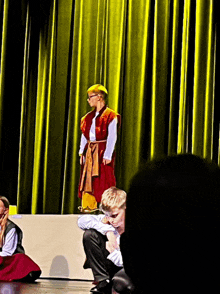 a boy in a red vest stands on a stage in front of a yellow curtain