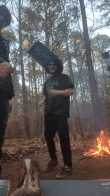 a man is standing in front of a fire in the woods with a book on his head .