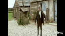 a man in a suit and hat is walking down a gravel road in front of a wooden building .