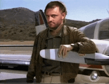 a man in a brown jacket leans on the wing of a plane