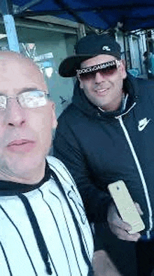 two men are standing next to each other and taking a selfie . one of the men is wearing sunglasses and a hat .