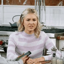 a woman in a purple and white striped sweater is sitting at a table in a kitchen holding a cell phone .